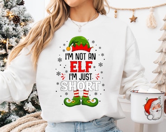 May include: White sweatshirt with the text "I'M NOT AN ELF I'M JUST SHORT" in red and black. The design includes an elf hat, red and green elf shoes, and snowflakes. A mug with a Santa Claus graphic is held in the image.