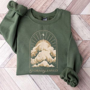 May include: Green sweatshirt with a graphic of a person reaching towards the sun above clouds, within an arched frame. The text "STORMBLESSED" is below the graphic. The sweatshirt has cuffed sleeves and a crew neck.