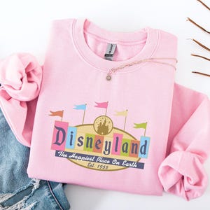 A pink sweatshirt with a Disneyland logo and text, along with a denim jacket and some leaves, is displayed on a white background.
