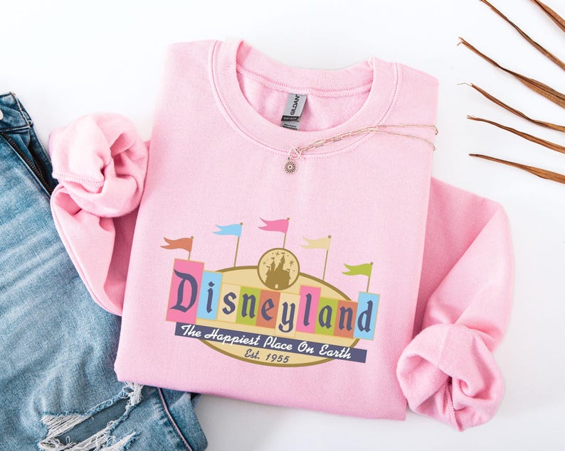 A pink sweatshirt with a Disneyland logo and text, along with a denim jacket and some leaves, is displayed on a white background.