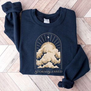 May include: Navy blue sweatshirt with a gold graphic of a person on a cloud, sun rays, and the text "STORMBLESSED". The sweatshirt is made of a soft material and has a crew neck and long sleeves.