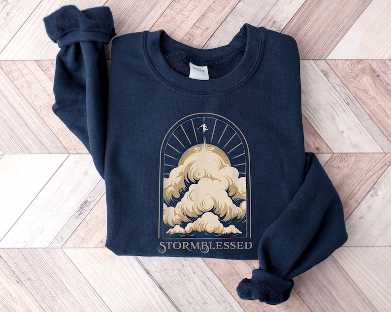 May include: Navy blue sweatshirt with a gold graphic of a person on a cloud, sun rays, and the text "STORMBLESSED". The sweatshirt is made of a soft material and has a crew neck and long sleeves.