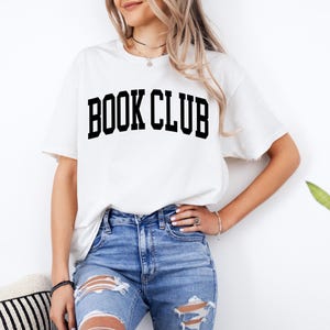 May include: White t-shirt with the text "BOOK CLUB" printed in black.