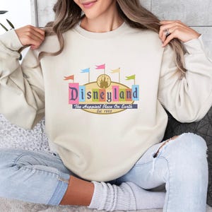 A woman wearing a white sweater with a colorful graphic of Disneyland on it, sitting on a couch.