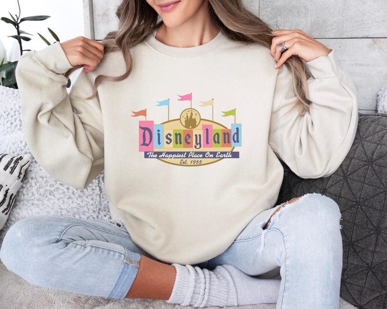 A woman wearing a white sweater with a colorful graphic of Disneyland on it, sitting on a couch.