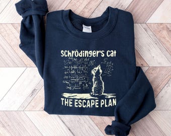 May include: Navy blue sweatshirt with the text "Schrödinger's Cat" and "The Escape Plan" in a white, handwritten-style font. A cat illustration is also featured. The sweatshirt is folded on a light wood surface.