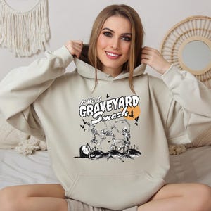 May include: Beige hoodie with a front graphic that reads "It Was A Graveyard Smash" in white and orange. The graphic features skeletons dancing in a graveyard scene with bats and a full moon. The hoodie has a kangaroo pocket and a drawstring hood.