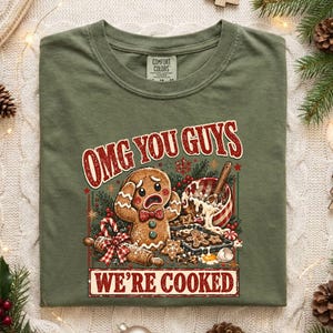 May include: An olive green t-shirt featuring a graphic of a distressed gingerbread person with the text "OMG YOU GUYS WE'RE COOKED" above and below. The illustration includes a rolling pin, a mixing bowl, and small gingerbread cookies.