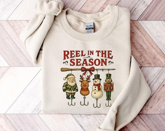 May include: A cream-colored sweatshirt with the text "REEL IN THE SEASON" in red and green. Below the text, a fishing rod holds Santa, a reindeer, a snowman, and a nutcracker, each hanging from a fishing hook.
