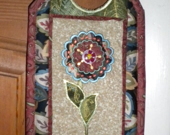 May include: A decorative door hanger with a floral design. The hanger is made of fabric and has a brown, green, and red color scheme. It features a large embroidered flower with a pink center and a gold-colored stem and leaves.