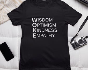 May include: Black t-shirt with the word "WOKE" in white capital letters, with the words "WISDOM, OPTIMISM, KINDNESS, EMPATHY" stacked vertically below. The shirt is on a white surface with a patterned design.