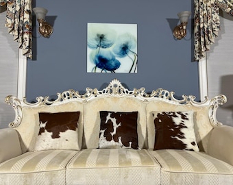 May include: A white, ornate sofa with three brown and white cowhide pillows. The sofa is in front of a wall with a blue and white floral print framed artwork.
