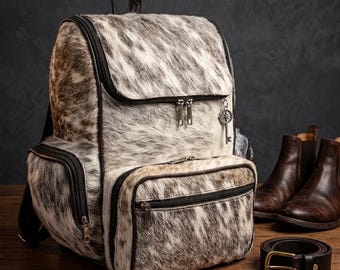 Puede incluir: Una mochila de piel de vaca con ribetes y cremalleras negras, un asa superior y un dije de llave. La bolsa está junto a un par de botas de cuero marrón y un cinturón de cuero negro con hebilla dorada.
