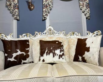 May include: A cream sofa with three cushions. The cushions have a brown and white cowhide pattern. The sofa has a detailed, ornate white frame with gold accents. The sofa is in front of a window with patterned curtains.