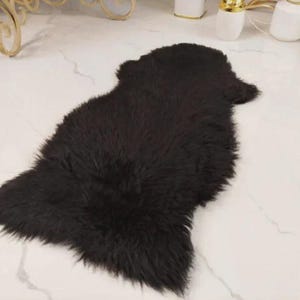 May include: A dark brown, fluffy sheepskin rug. The rug is irregularly shaped with a dense, shaggy texture. It is placed on a white marble-patterned floor, with decorative items visible in the background.