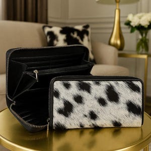 May include: Two black leather wallets, one open and one closed, sit on a gold table. The closed wallet has a white and black spotted pattern. The open wallet reveals multiple compartments. The background includes a sofa, a pillow with a similar pattern, and a gold lamp.