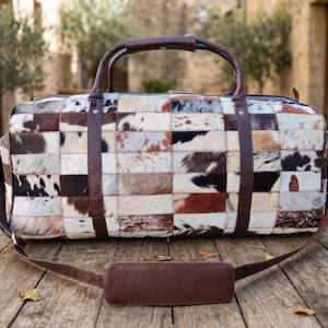 May include: A patchwork leather duffel bag with a brown leather handle and shoulder strap. The bag is made of various rectangular pieces of cowhide in shades of brown, white, and black. The bag is sitting on a wooden surface.