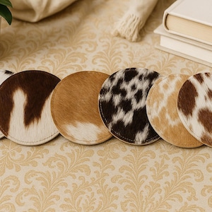 May include: A set of six round coasters with cowhide patterns in various colors, including black and white, brown, and tan. The coasters are arranged on a patterned surface, suggesting a home decor theme.