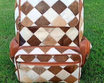Op de afbeelding: Een rugzak met een patchwork-ontwerp met bruine, witte en bruinachtige ruitvormen. De rugzak heeft bruine leren accenten, een voorvak en zijvakken. De tas staat op groen gras.