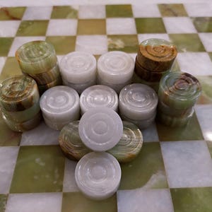 May include: A checkerboard with green and white squares, holding a collection of checkers. The checkers are stacked, showcasing a mix of white and green-brown colours. The game pieces are made of a stone-like material.