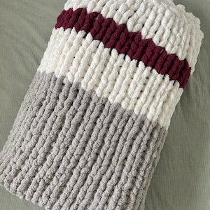 Könnte beinhalten: Eine dicke Strickdecke mit Streifen aus weißem, burgunderrotem und grauem Garn.