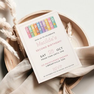 Può includere: Un biglietto d'invito rosa chiaro per il secondo compleanno di Matilda, con illustrazioni di libri colorati e la scritta "CHAPTER TWO". Il biglietto include la data, l'ora e il luogo della festa, appoggiato su un vassoio di legno con elementi decorativi.