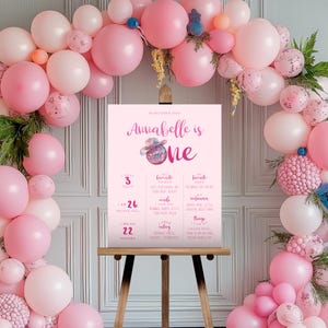 May include: A pink and white printable first birthday sign with a watercolor design. The sign includes the name Annabelle, the number one, and details about the child's age, height, weight, and favorite things. The sign is displayed on a wooden easel in front of a pink balloon arch.