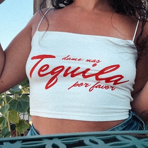 Puede incluir: Top blanco de tirantes con el texto "dame mas Tequila por favor" en letras rojas.