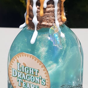May include: A small, clear glass bottle with a teal blue liquid inside. The bottle has a gold and white wax seal on the top and a brown twine tied around the neck. The bottle has a label that says "Light Dragon's Tears".