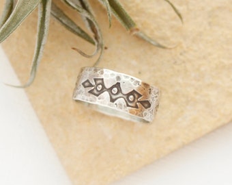 May include: A silver band ring with a hammered texture and a geometric design. The ring is on a light brown surface.