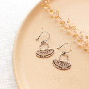 May include: A pair of silver earrings with a brass, fan-shaped design. The earrings have a textured, geometric pattern and are hanging on a beige surface.