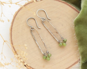 May include: A pair of handcrafted silver earrings. Each earring features a long, hammered silver oval with a cluster of small, round, light green beads at the bottom. The earrings are displayed on a wooden surface with a green fabric in the background.
