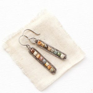 May include: A pair of handcrafted earrings featuring a rectangular silver frame filled with colorful, square-shaped beads. The beads are in shades of brown, beige, green, and orange. The earrings have a hook closure and are displayed on a cream-colored cloth.