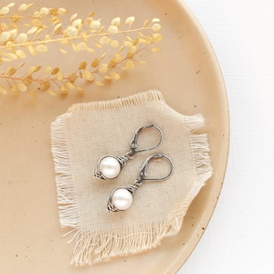 May include: A pair of silver earrings with a pearl wrapped in wire, displayed on a beige fabric surface.