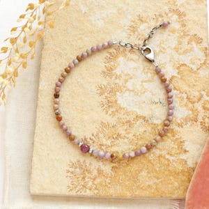 Puede incluir: Una delicada pulsera con pequeñas cuentas redondas de piedras preciosas rosas y marrones. La pulsera tiene una cadena y un cierre de color plateado. Una gema rosa más grande y facetada está engastada en el centro. La pulsera se muestra sobre una superficie clara y texturizada.