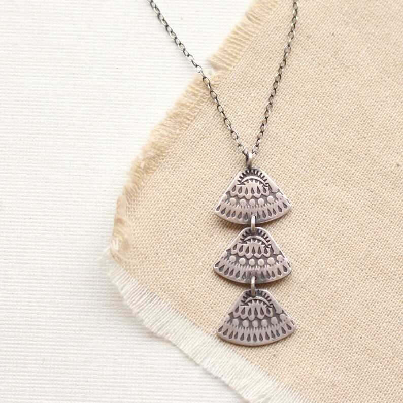 Pode incluir: Um colar de prata com um pingente de tr&ecirc;s n&iacute;veis. Cada n&iacute;vel &eacute; um tri&acirc;ngulo de prata com um padr&atilde;o intrincado em relevo.