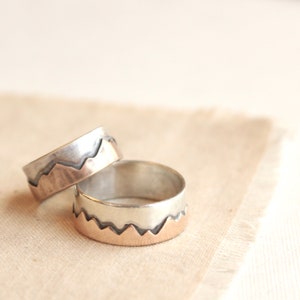 May include: Two silver rings with a mountain range design. The top of the mountains are a rose gold color.