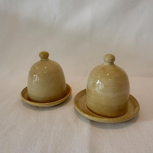 May include: Two matching ceramic butter dishes with dome-shaped covers and small knob handles. Each piece features a smooth, glossy, light beige glaze with subtle tonal variations and sits on a circular base plate with a slightly raised rim.