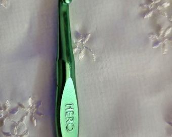 Può includere: Un uncinetto in metallo verde con un fusto liscio e affusolato e una testa curva. La parola HERO è impressa in lettere maiuscole sull'area piatta del poggiapollice del manico, che presenta una finitura lucida e una forma cilindrica.