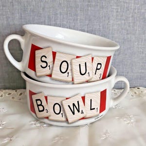 May include: Two white ceramic soup bowls with handles, stacked vertically. Each bowl features a red horizontal band decorated with beige tiles displaying the letters S, O, U, P on the top bowl and B, O, W, L on the bottom bowl, mimicking a word game.