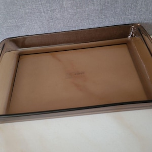 Könnte beinhalten: Eine rechteckige Auflaufform aus durchscheinendem braunem Glas. Die Form hat eine glatte, polierte Oberfläche mit abgerundeten Ecken und einem breiten, flachen Rand. In der Mitte des Bodens befinden sich schwache, kaum sichtbare Prägungen.