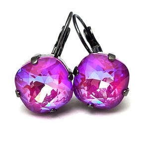 Puede incluir: Un par de pendientes colgantes con un cristal morado iridiscente en cada pendiente. Los pendientes tienen un gancho de metal negro.