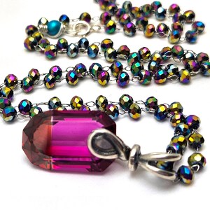 Op de afbeelding: Een ketting met een grote, gefacetteerde, roze kristallen hanger. De ketting is gemaakt van iriserende zwarte kralen.