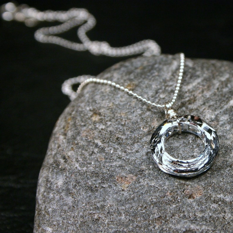 May include: A silver chain necklace with a clear crystal pendant in the shape of a circle. The pendant is made of faceted crystal and has a wire wrapped around the edge.
