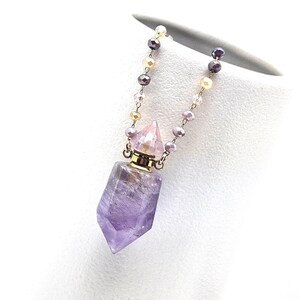 May include: A purple amethyst crystal pendant necklace with a gold-colored metal cap and a beaded chain. The chain features small, round beads in shades of purple, pink, and gold. The pendant is a faceted, elongated crystal shape.