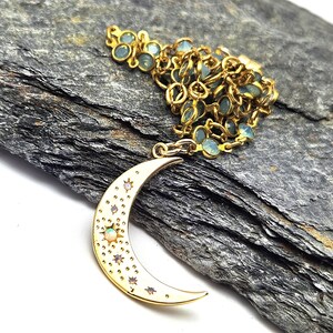 May include: Gold-toned necklace featuring a crescent moon pendant with a white enamel finish and embedded star and opal accents. The necklace has a delicate chain with small, light blue gemstone beads.
