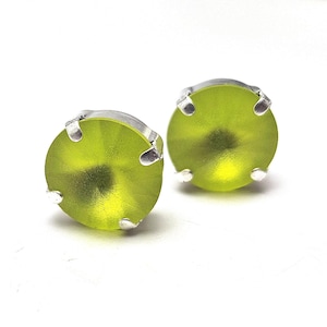May include: A pair of round, lime green stud earrings. Each earring features a vibrant, translucent green stone set in a silver-colored metal bezel with four prongs. The earrings are displayed against a white background.