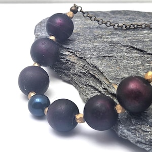 May include: A necklace featuring large, dark purple beads interspersed with smaller gold-colored beads and a single dark blue bead. The necklace is set against a textured gray stone background, with a delicate chain.