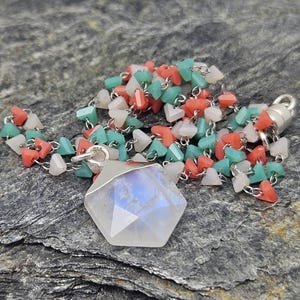 May include: A necklace with a large, faceted, hexagonal moonstone pendant. The chain is composed of small, colorful beads in coral, mint green, and white. The necklace has a silver clasp and is set against a textured gray stone surface.