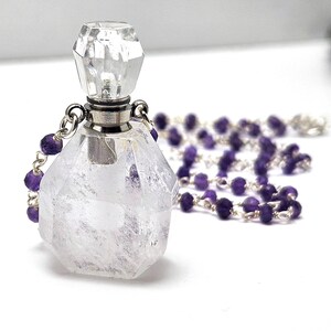 May include: A clear, faceted crystal perfume bottle with a silver-colored cap and a matching crystal stopper. A strand of purple amethyst beads is attached to the bottle. The bottle is on a white surface.